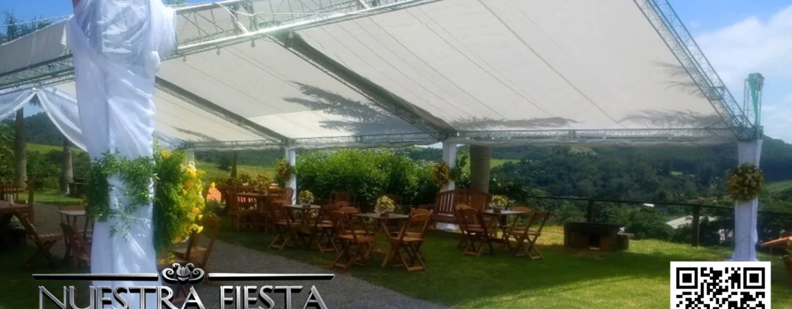 Tenda grande da NUESTRA FIESTA decorada com tecido branco e flores na estrutura, mesas e cadeiras de madeira embaixo, com montanhas verdes ao fundo em um dia ensolarado