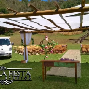 Cenário de casamento montado em um gramado, com cadeiras de madeira da NUESTRA FIESTA dispostas ao redor de um pergolado rústico de madeira decorado com tecidos brancos.