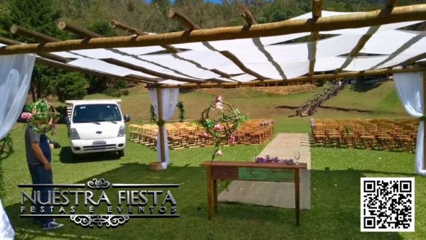 Cenário de casamento montado em um gramado, com cadeiras de madeira da NUESTRA FIESTA dispostas ao redor de um pergolado rústico de madeira decorado com tecidos brancos.